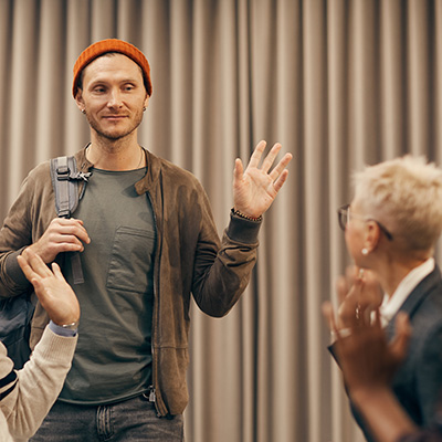 Guy wearing a beanie and backpack waving to the group as he enters the room